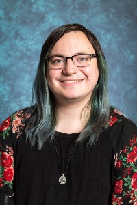 Picture of Lore Falls, Program Coordinator of LGBT+ Programs  The image portrays a smiling individual with straight, shoulder-length hair that is dark at the roots and transitions into a shade of teal at the ends. She is wearing rectangular eyeglasses with black frames. She is dressed in a black top, which features sleeves adorned with a pattern of red and pink flowers and green leaves. Around her neck, she wears a black cord necklace with a round pendant that has a design in the center. The background is an evenly lit, textured blue backdrop, adding a neutral, non-distracting context to the portrait.